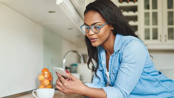 Young African woman at home, leaning on her kitchen counter, texting on her phone while enjoying coffee.