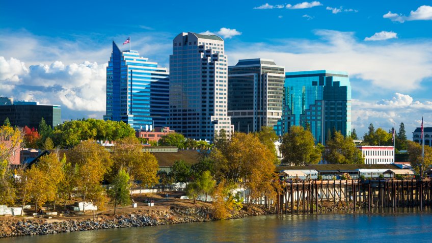 Sacramento downtown skyline during Autumn with Autumn colored trees in the foreground, with the Sacramento River, and dramatic clouds in the background.