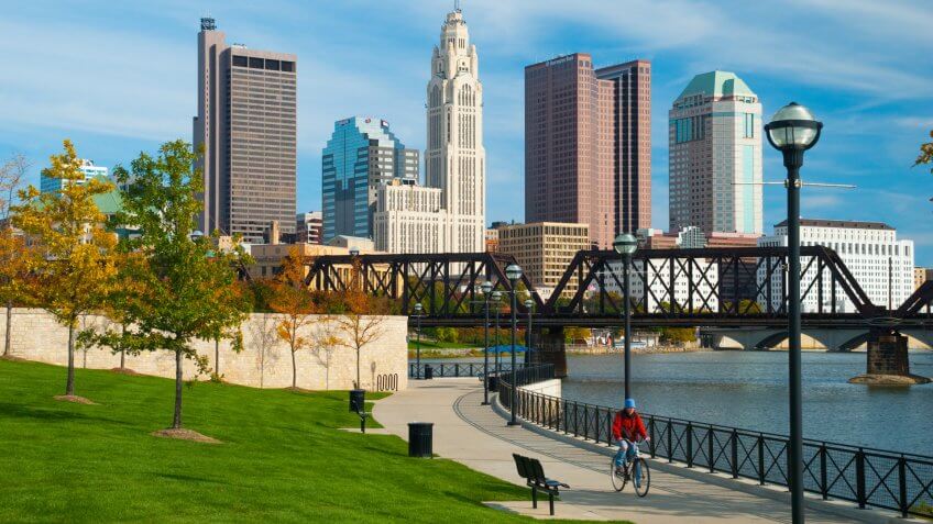 "Columbus, United States - October 19, 2009: A person riding a bike on a concrete path beside the Scioto River with trees and a train bridge in the foreground and the Columbus downtown skyline in the background.