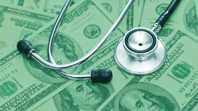 A stethoscope lays on top of a stack of $100 bills, which is covered in a green background