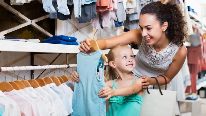 Positive young mother with daughter buying kids clothes in blue color in shop.
