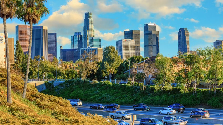 Palm trees and freeway traffic fills the foreground leading back to the skyscrapers of Los Angeles skyline with cloudscape above, California.