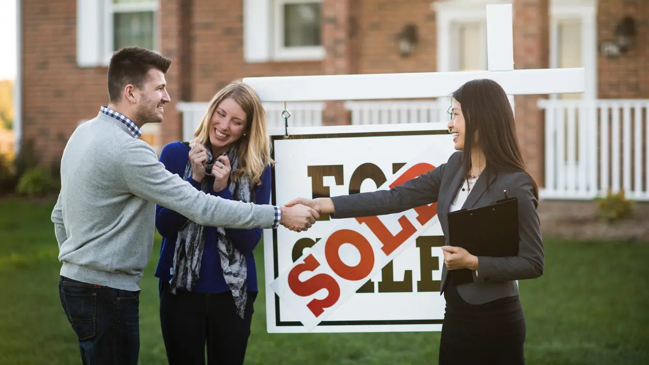 happy couple standing next to for sale sign shaking hands with real estate agent
