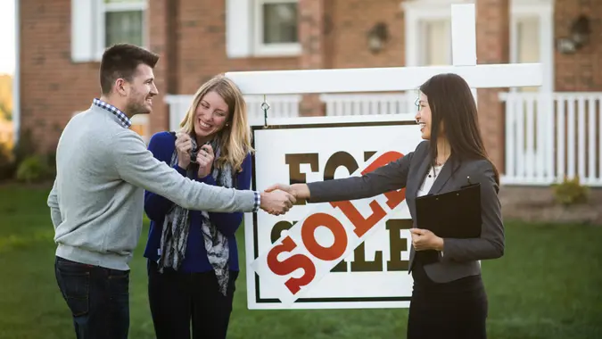 happy couple standing next to for sale sign shaking hands with real estate agent