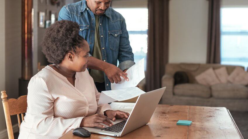 Smiling African couple paying bills and doing their online banking with a laptop at their kitchen table at home.
