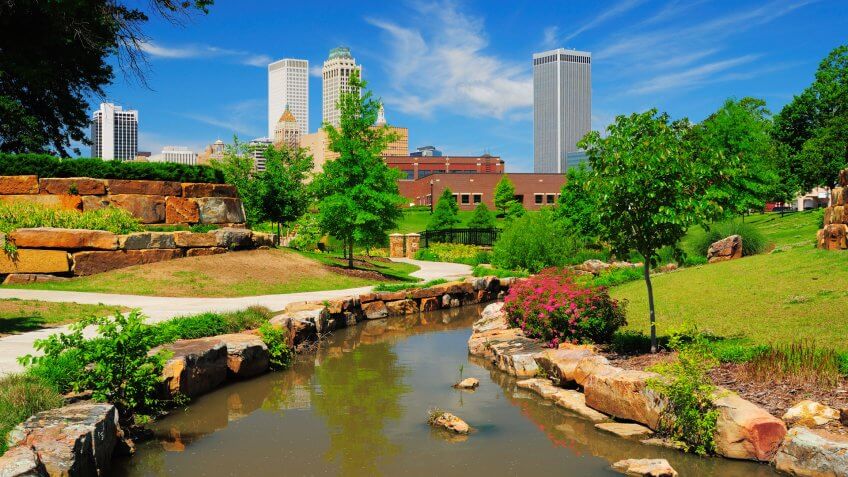 "Tulsa downtown skyline from a park with trees, grass, rocks, and a stream in the foreground.