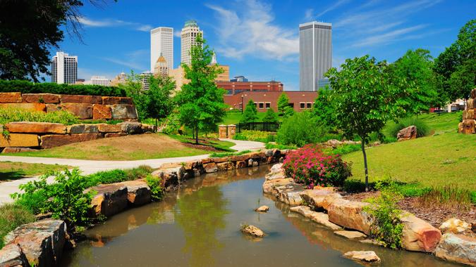 "Tulsa downtown skyline from a park with trees, grass, rocks, and a stream in the foreground.