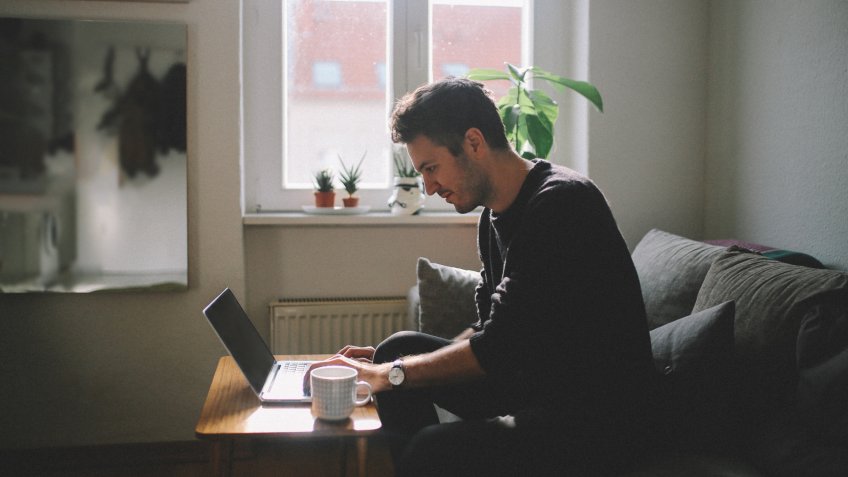 Vintage tone portrait image of a young man working at home at the laptop, sitting at the wooden table, having a cup of coffee.