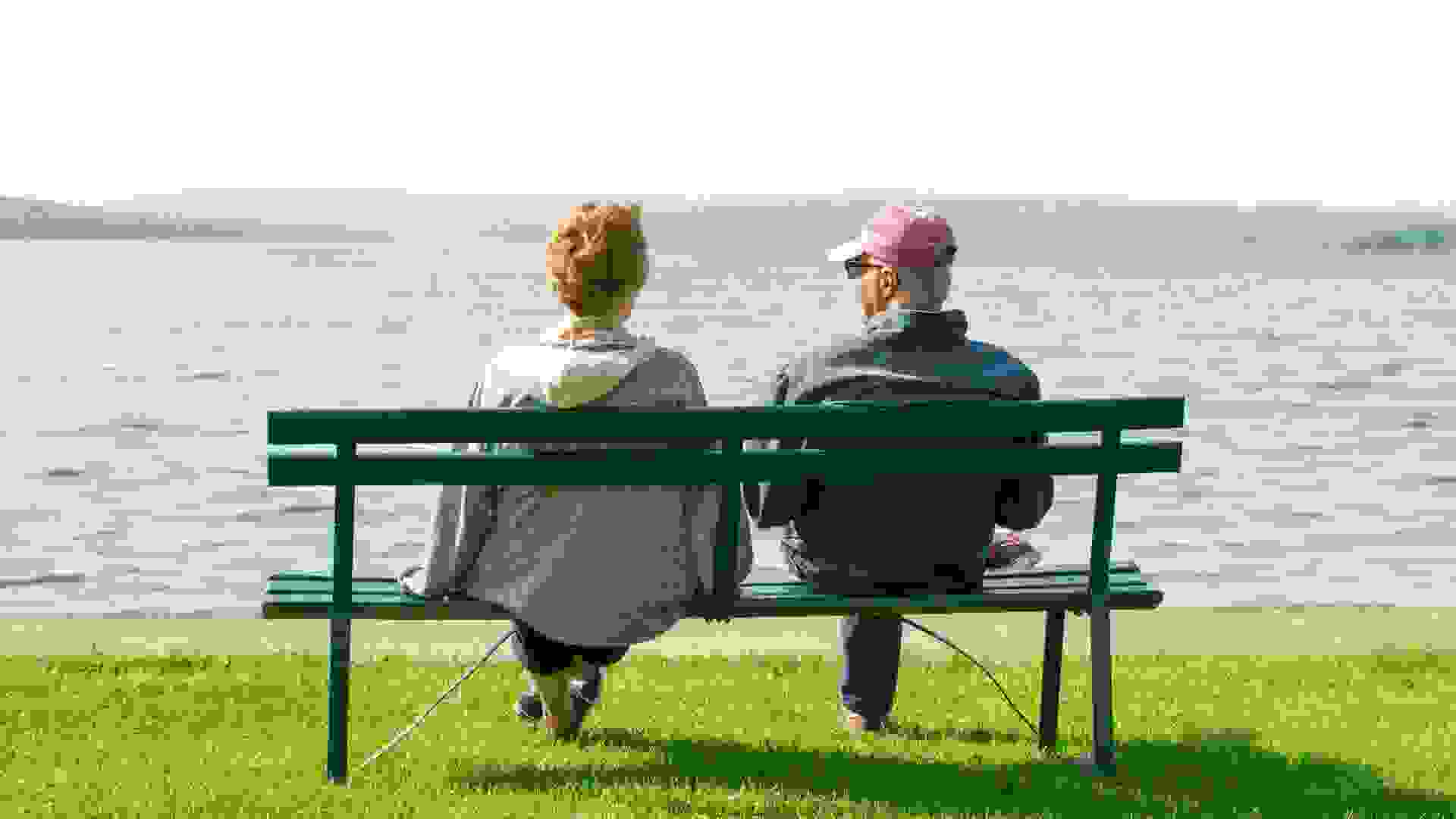 An older elderly retired couple sit on a park bench casually dressed looking at a view of a beautiful lake.