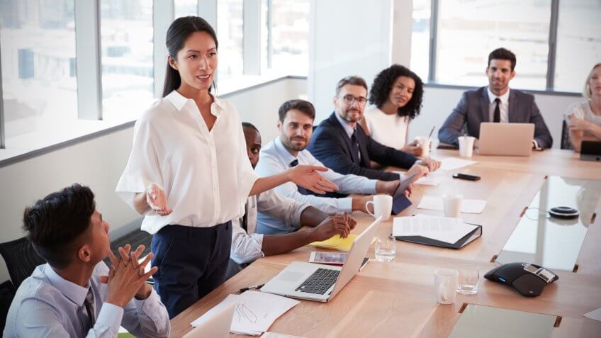 Businesswoman Stands To Address Meeting Around Board Table.
