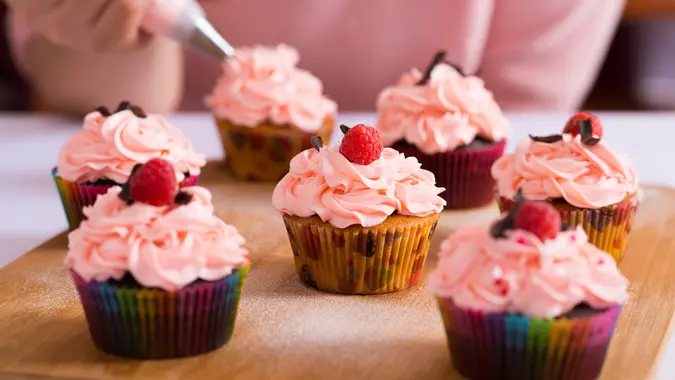 Close shot of many sweet cupcakes on the foreground while a baker decorating the last one.