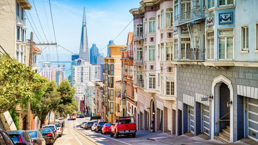 San Francisco, USA - May 10, 2016: Pacific heights street scene panoramic downhill view towards downtown with parked cars, a tramway, a man waiting in a doorway and the Transamerica pyramid in the background.