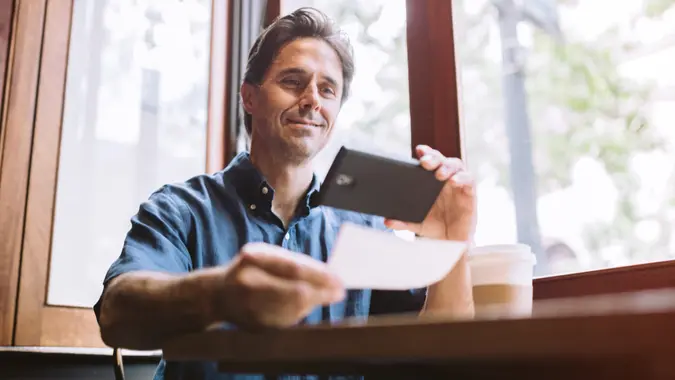 A mature smiling man in his mid 40's takes a picture with his smart phone of a check or paycheck for digital electronic depositing, also known as "Remote Deposit Capture", fees, America, money, payment, avoid fees, bills, debt