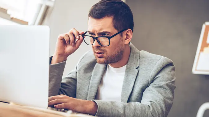 Handsome young man looking at his laptop and adjusting his glasses while sitting at his working place, fees, America, money, payment, avoid fees, bills, debt