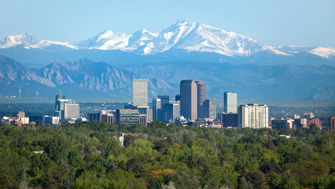 Snow covered Longs Peak, part of the Rocky Mountains stands tall in the background with green trees and the Downtown Denver skyscrapers as well as hotels, office buildings and apartment buildings filling the skyline.