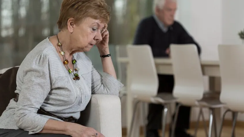 Upset elderly woman sitting on couch with husband in background