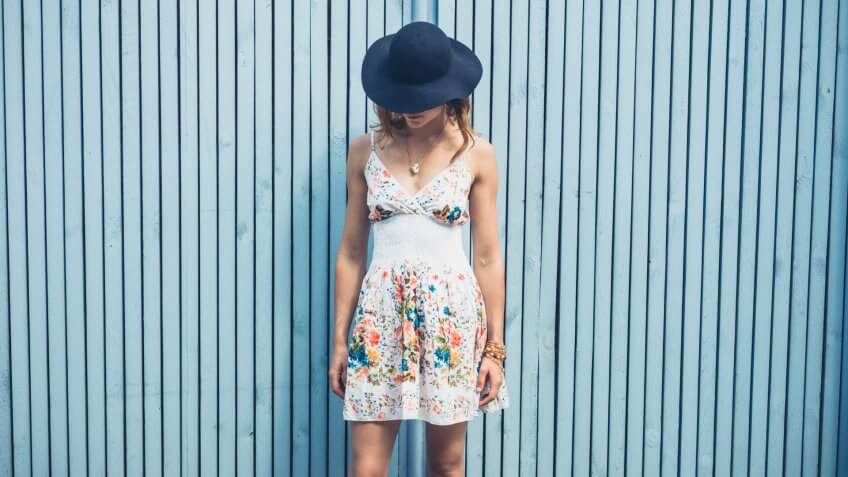 A young woman wearing a summer dress with a hat is standing outside by a blue fence