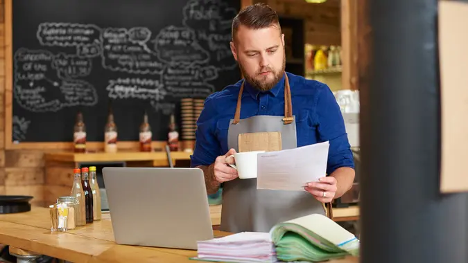 a coffee shop owner checks the delivery notes in his bookkeeping folder on the counter of his busy coffee shop and cross checks them with his online accountancy via his laptop at the end of a busy day .