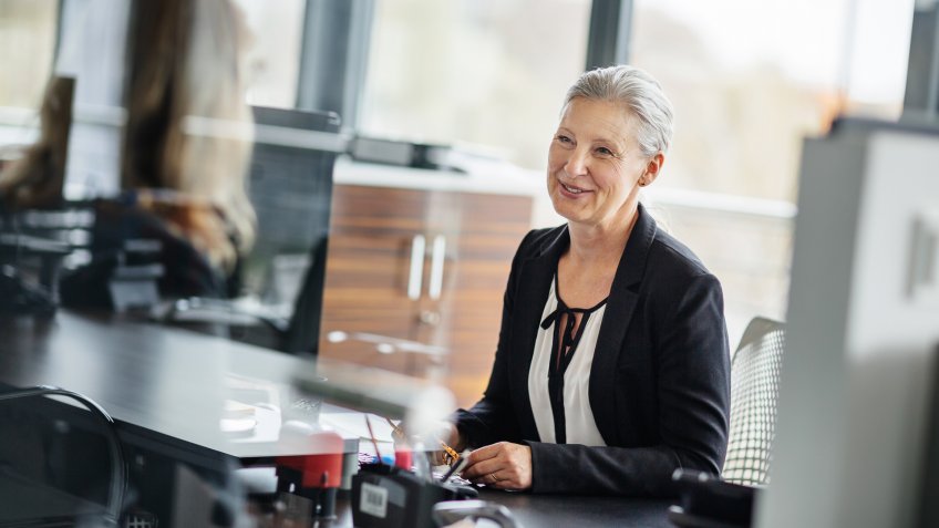 Senior businesswoman sitting on her desk in a modern office and talking with an employee.