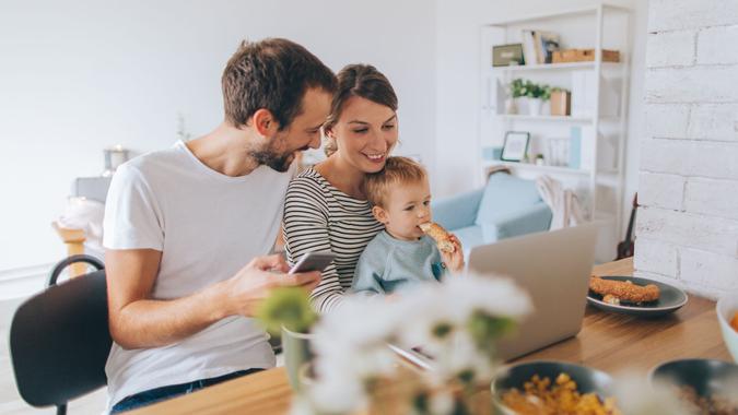 Photo of a young family is showing their hectic lifestyle - busy morning they are spending together over dinning table, using computer and mobile phone while their toddler boy is with them.