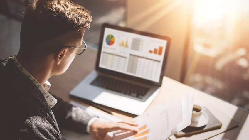 Young finance market analyst in eyeglasses working at sunny office on laptop while sitting at wooden table. Young finance market analyst in eyeglasses working at sunny office on laptop while sitting at wooden table.