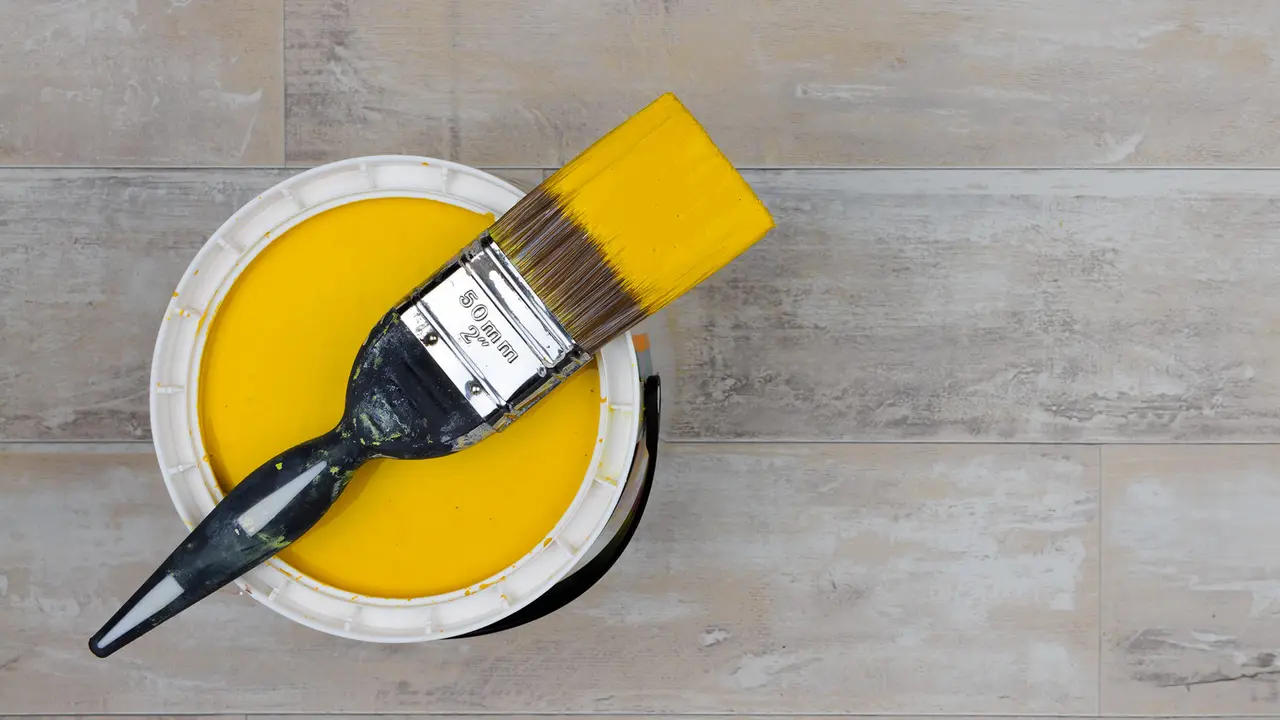 Looking down on a can of Bright Yellow Paint with a loaded brush stood on a shabby style wood floor.
