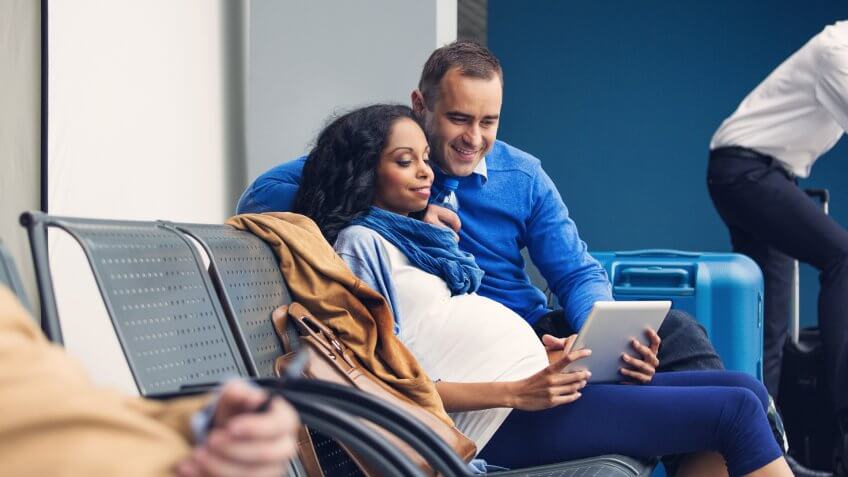 Pregnant woman waiting with her partner for the flight at the airport lounge, using a digital tablet together.