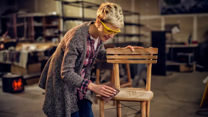 Attractive satisfied female furniture designer carefully sanding a chair frame with shelves of wooden items behind her in the workshop.
