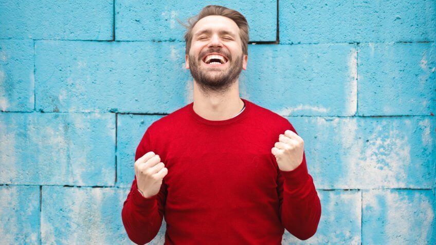 Smiling man in red sweater against blue wall making happy victorious gesture
