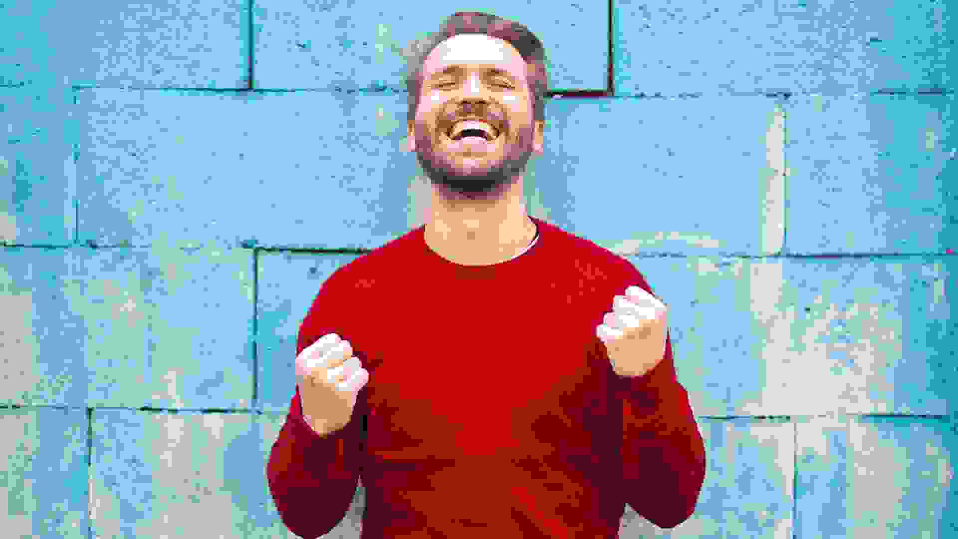 Smiling man in red sweater against blue wall making happy victorious gesture