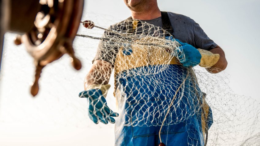 Part of fisherman's body, wearing yellow and blue pants and standing on a fishing boat while pulling the fishing nets in Koper, Slovenia.