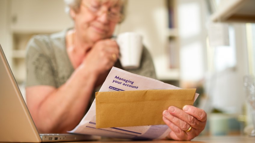 Senior woman looking at a bank statement at her laptop in the kitchen.
