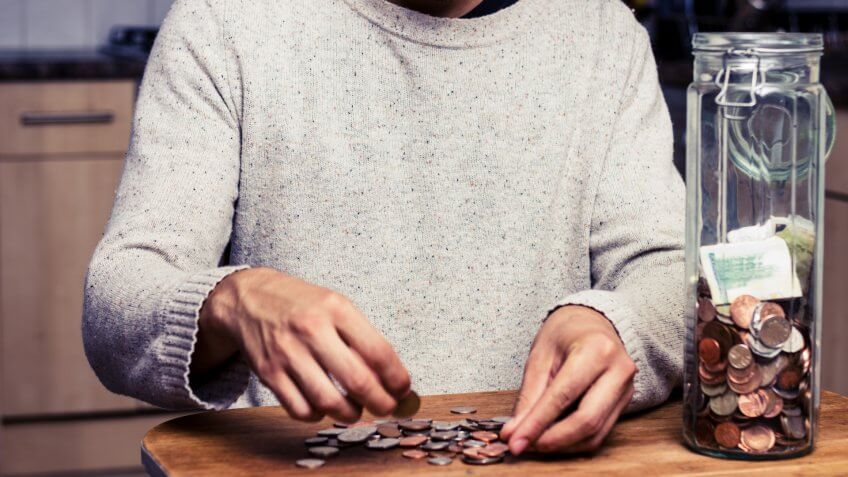 Young man is sitting at a table in his kitchen and counting the money from his piggy bank, a tall glass jar.