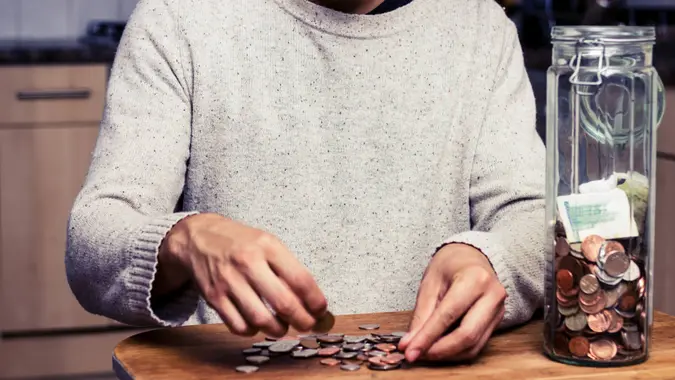 Young man is sitting at a table in his kitchen and counting the money from his piggy bank, a tall glass jar.