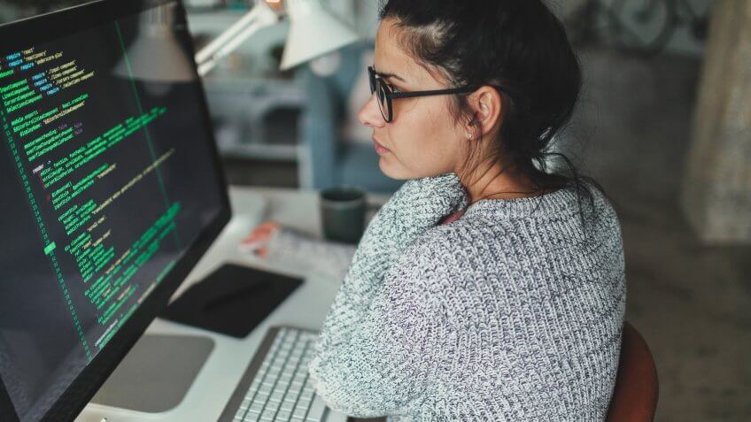 Photo of a young woman sitting at her desktop computer, doing computer programming in her home office.