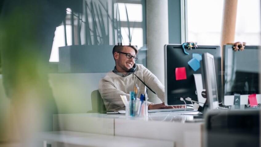 Business professional typing notes with keyboard on computer while talking and smiling on the phone to client.