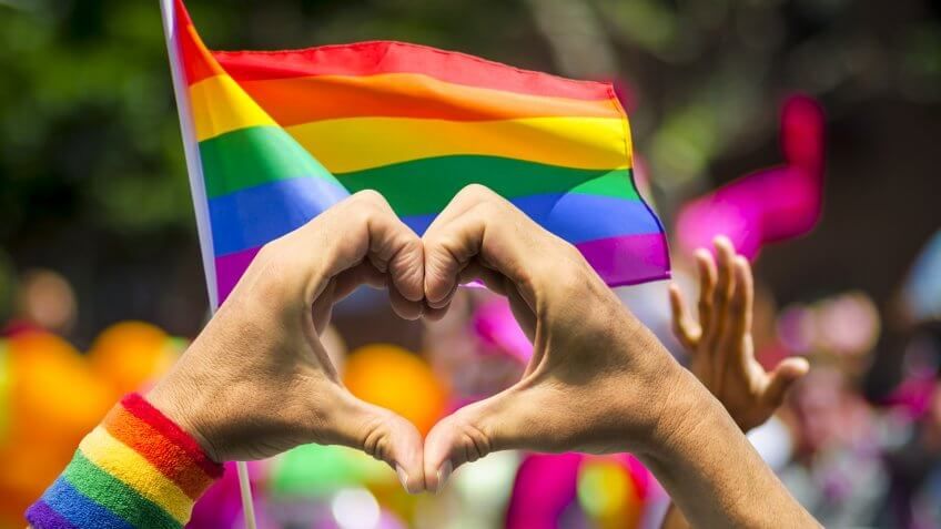Supporting hands make heart sign and wave in front of a rainbow flag flying on the sidelines of a summer gay pride parade.