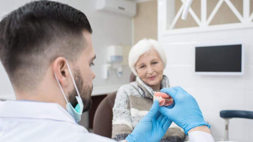 Professional male dentist in gloves showing dentures teeth to senior female patient and explaining how to use it in dental surgery.
