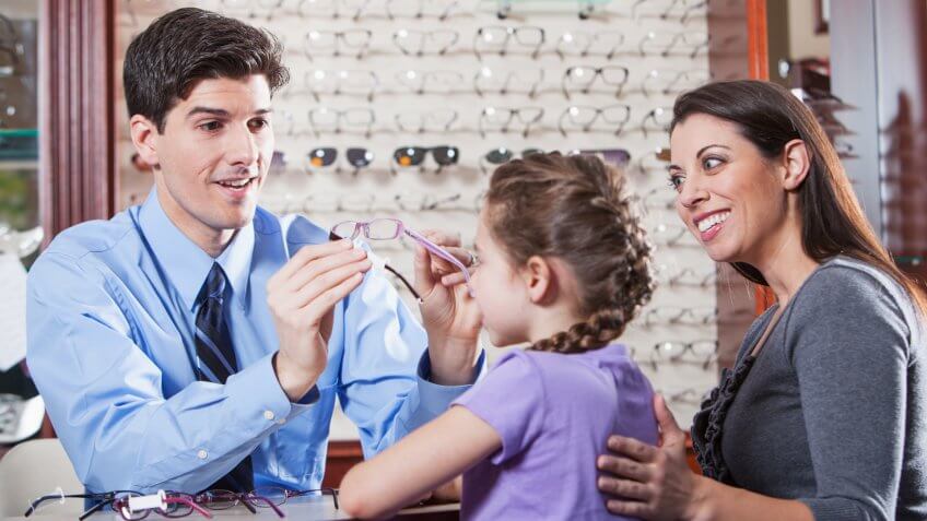 Optician helping little girl try on new glasses while mother watches