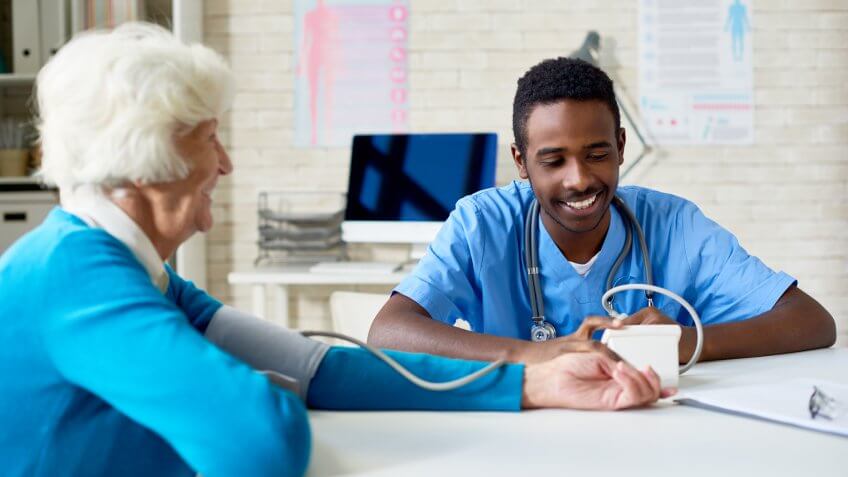 Smiling licensed vocational nurse working with senior patient and measuring blood pressure in the doctors office