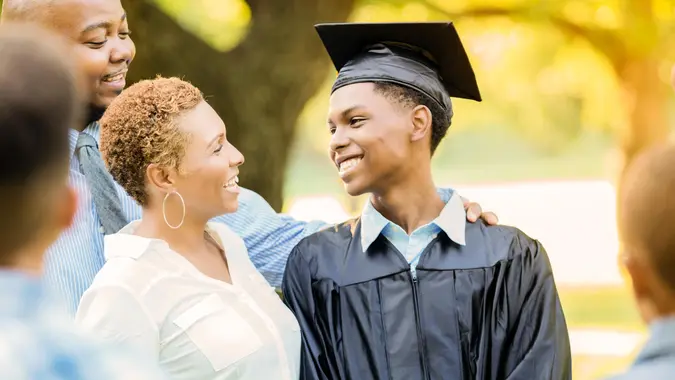 A young male high school graduate in cap and gown smiles at his proud mom and other family members.