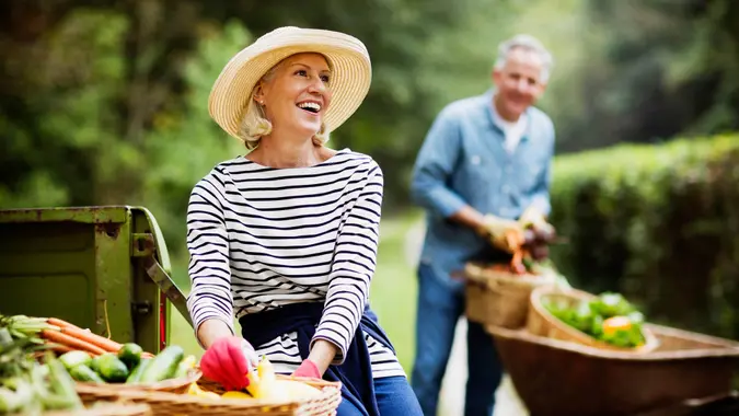 Portrait of mature woman with baskets of harvested vegetables and man working in the background.