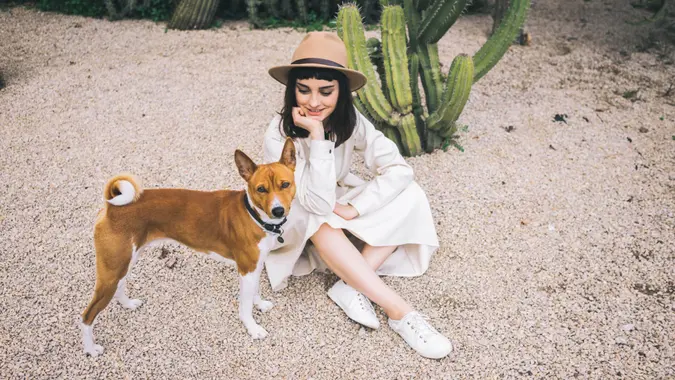 Happy brunette girl walks in cacti park in old fashioned white dress and brown hat with her clever basenji dog.