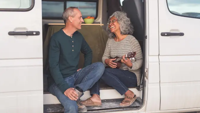 Senior couple sit in the doorway to their camper van while taking a break from driving. The couple is on a road trip in celebration of recently retiring. The ethnic woman strums a ukulele while singing her husband a song. Her husband is affectionately looking at her. He is holding a coffee cup. A portion of the interior of the van is visible. It is cloudy outside. The couple is dressed in casual clothing and the woman is wearing glasses.