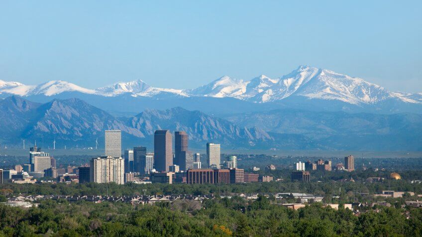 Snow covered Longs Peak, part of the Rocky Mountains stands tall in the background with the Downtown Denver skyscrapers as well as hotels, office buildings and apartment buildings filling the skyline.