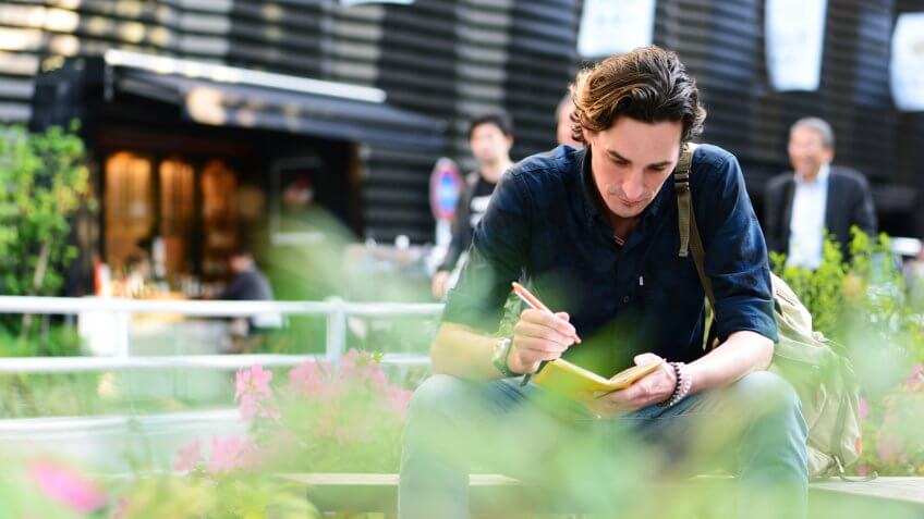 A mid adult man sitting outside in the late afternoon, writing something on a notebook.