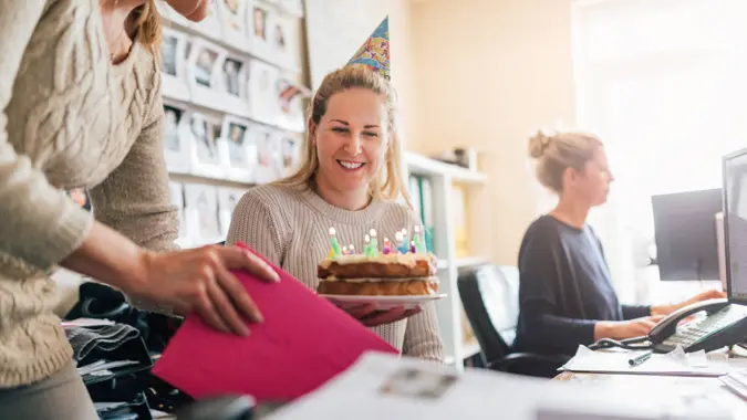 woman sitting at her office desk with a birthday cake handed to her by another female. She is wearing a party hat and smiling.