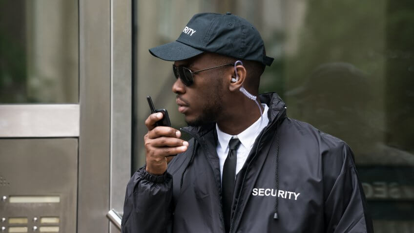 Security guard wearing sunglasses standing in front of entrance speaking into walkie talkie