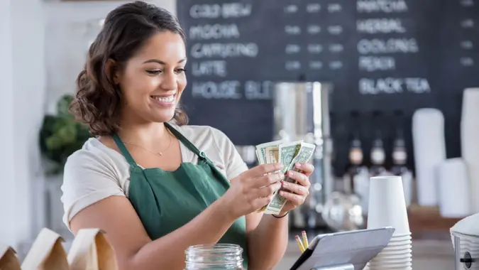 A smiling young female barista stands at the counter in her coffee shop and looks down as she counts paper bills that she has just pulled from her tips jar.