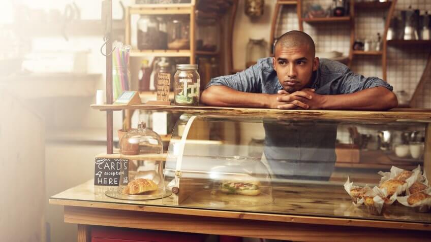 Shot of a young man standing behind the counter of his store and looking downhearted.